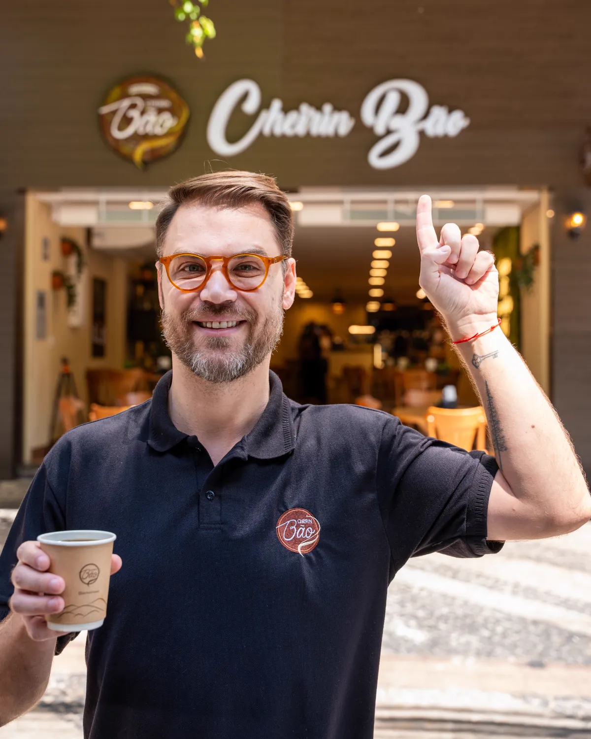 André, proprietário da Cheirin Bão Anchieta, com um café na mão apontando para a logo na fachada da loja