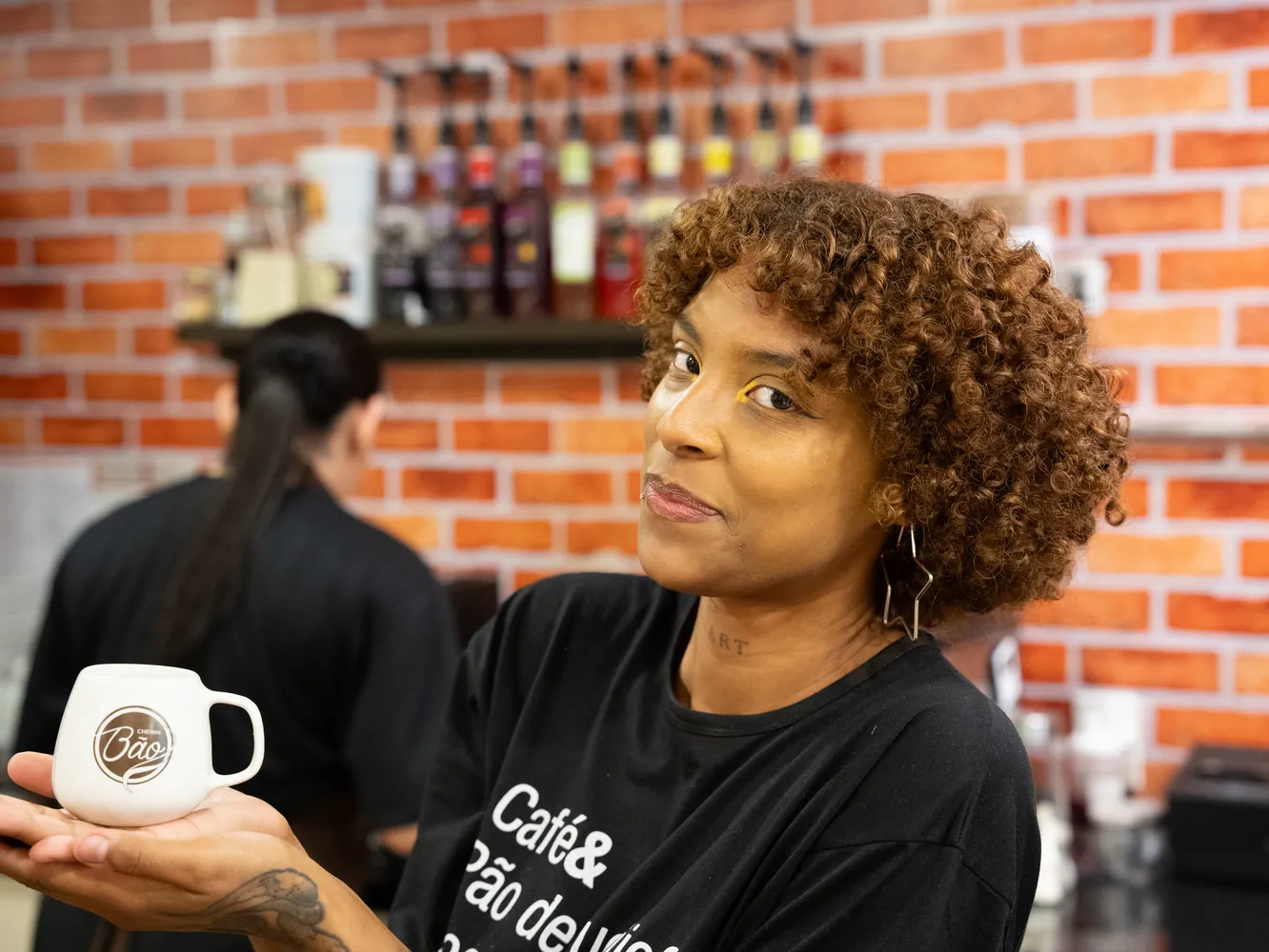 Atendente da Cheirin Bão Anchieta segurando a caneca Bão no balcão de tijolinho